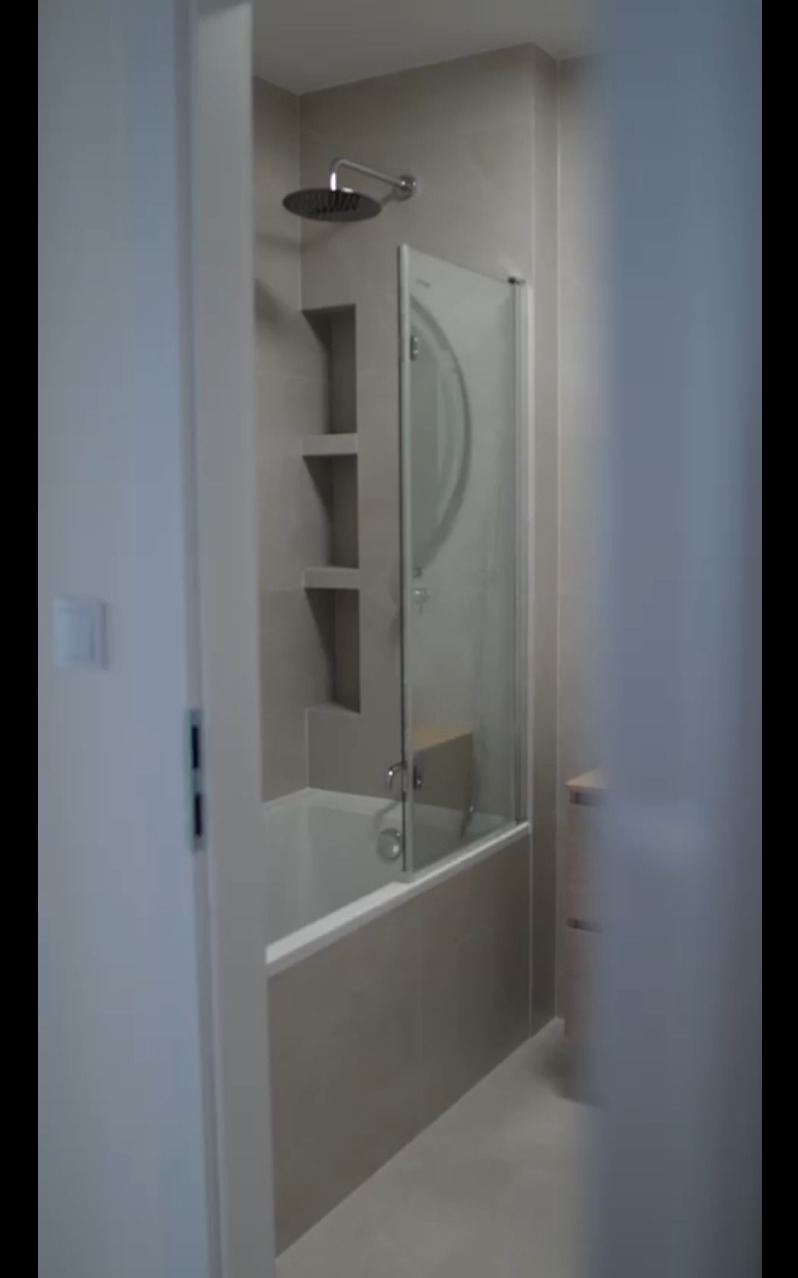 Nowoczesna łazienka z wanną i szklanym parawanem, wykończona w jasnych, neutralnych odcieniach szarości. Widoczna deszczownica i wnęki w ścianie.