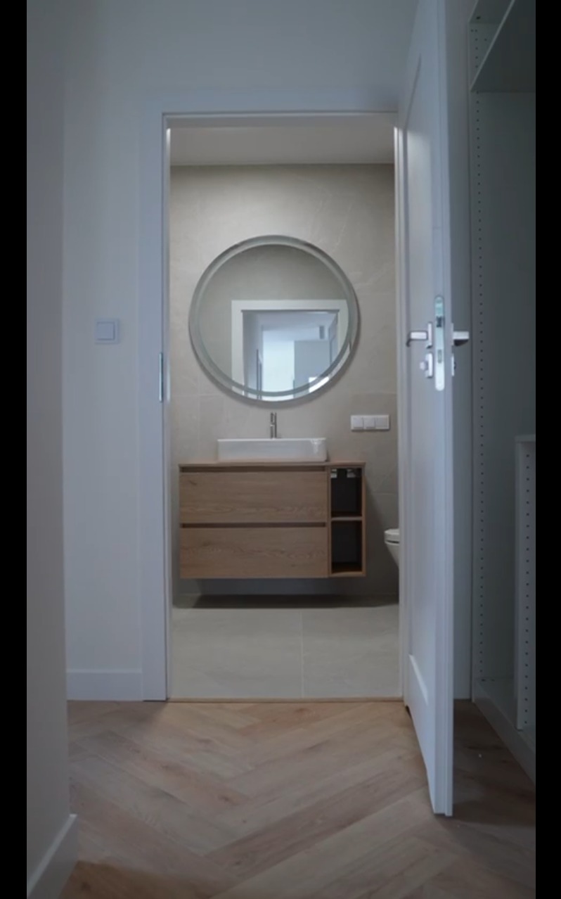 Nowoczesna łazienka z drewnianą szafką pod umywalką i okrągłym lustrem, widoczna z korytarza przez otwarte drzwi. Parkiet w jodełkę na podłodze.