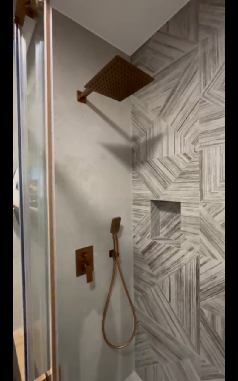 Nowoczesna kabina prysznicowa z geometryczną mozaiką na ścianie, deszczownicą i baterią w kolorze miedzi. Widoczny fragment szklanych drzwi.