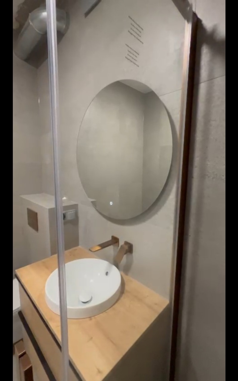 Nowoczesna łazienka z okrągłym lustrem podświetlanym LED, drewnianą szafką pod umywalką i złotą armaturą. Minimalistyczny design, szare płytki na ścianach.