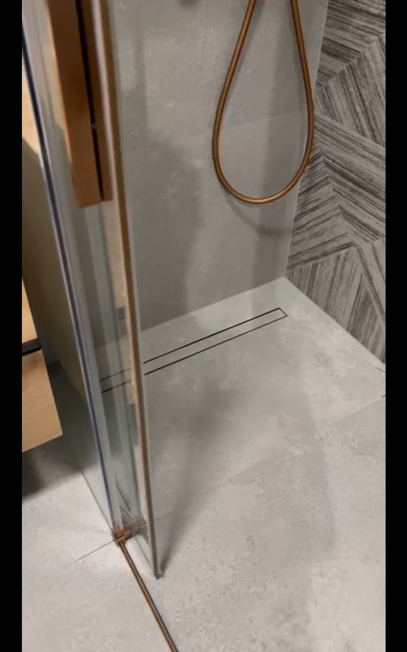 Nowoczesna kabina prysznicowa z odpływem liniowym i złotymi detalami, na tle szarej ściany z geometrycznym wzorem. Minimalistyczny design.