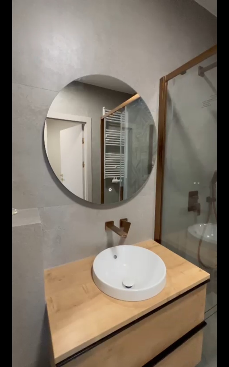 Nowoczesna łazienka po remoncie w Zielonej Górze: okrągłe lustro, drewniana szafka pod umywalką, kabina prysznicowa z brązowymi ramami i szarymi płytkami.