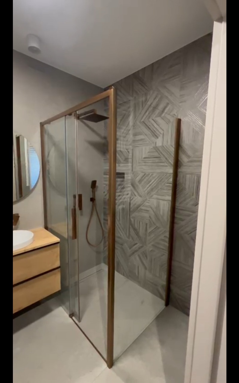 Nowoczesna kabina prysznicowa z brązowymi profilami i deszczownicą, umywalka z szafką w łazience z szarą, wzorzystą ścianą. Minimalistyczny design.
