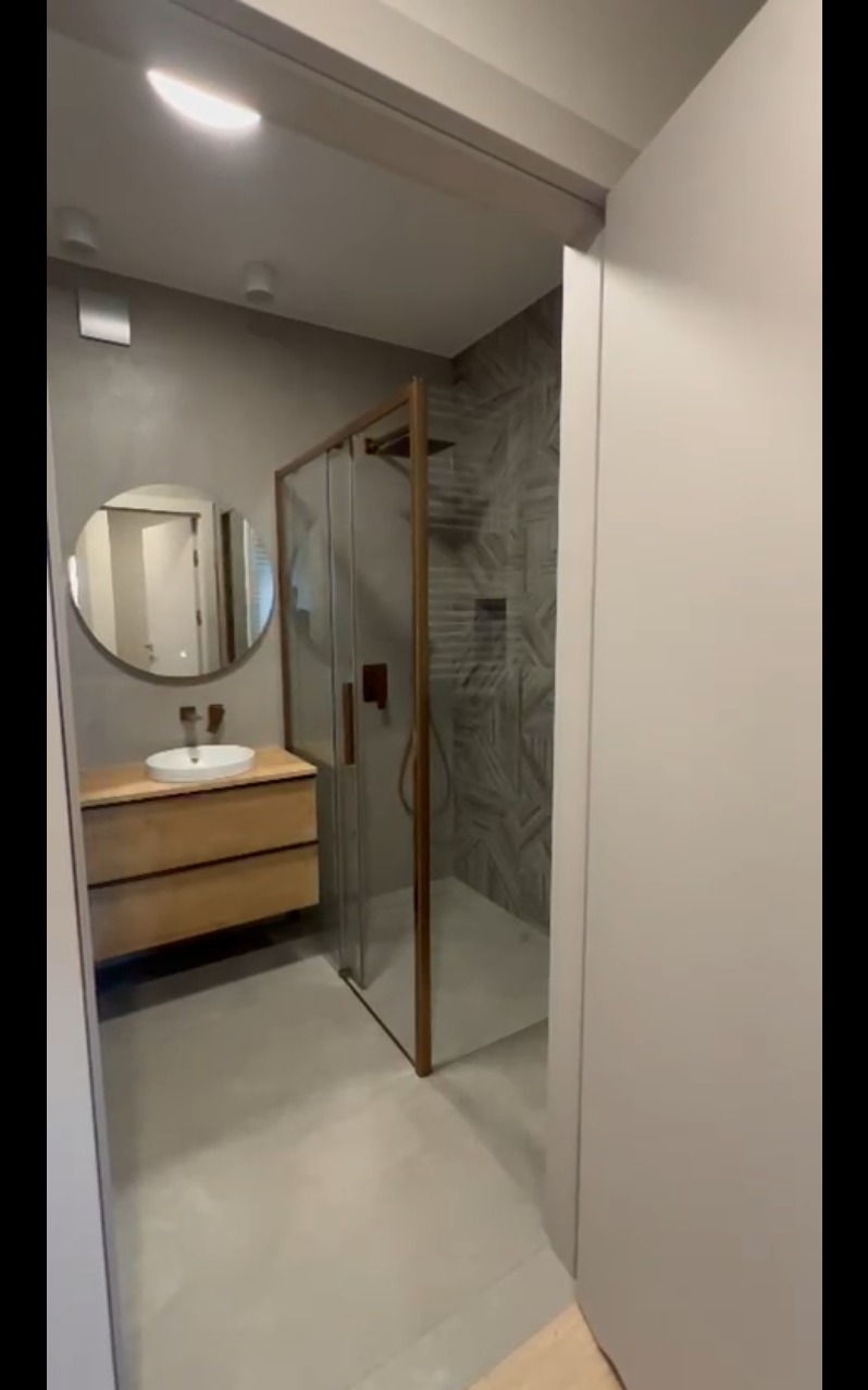 Nowoczesna łazienka z kabiną prysznicową w brązowej ramie, drewnianą szafką pod umywalką i okrągłym lustrem. Szare ściany i podłoga tworzą spójną całość.