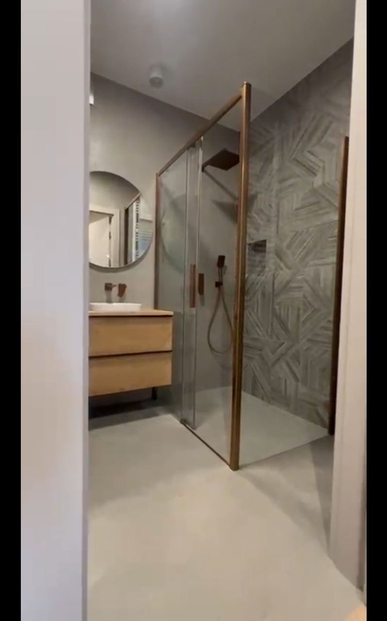 Nowoczesna łazienka z kabiną prysznicową w złotej ramie, umywalką na drewnianej szafce i okrągłym lustrem. Ściana wykończona płytkami w geometryczny wzór.