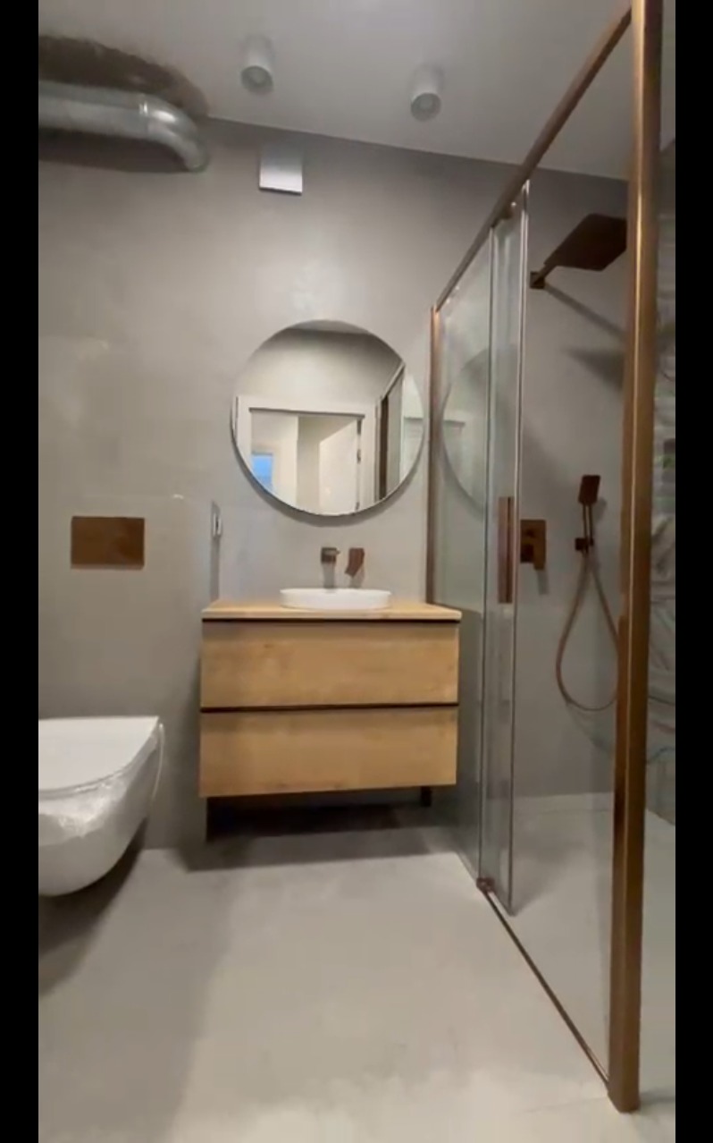 Nowoczesna łazienka z kabiną prysznicową typu walk-in, okrągłym lustrem nad drewnianą szafką umywalkową i minimalistyczną toaletą. Szare ściany i podłoga.