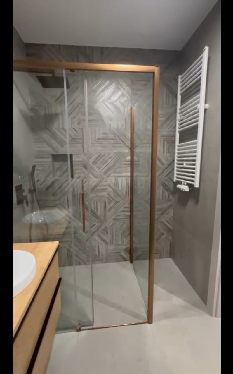 Nowoczesna kabina prysznicowa z geometryczną mozaiką na ścianie, złote ramy, biały grzejnik drabinkowy i umywalka z drewnianą szafką w wykończonej łazience.