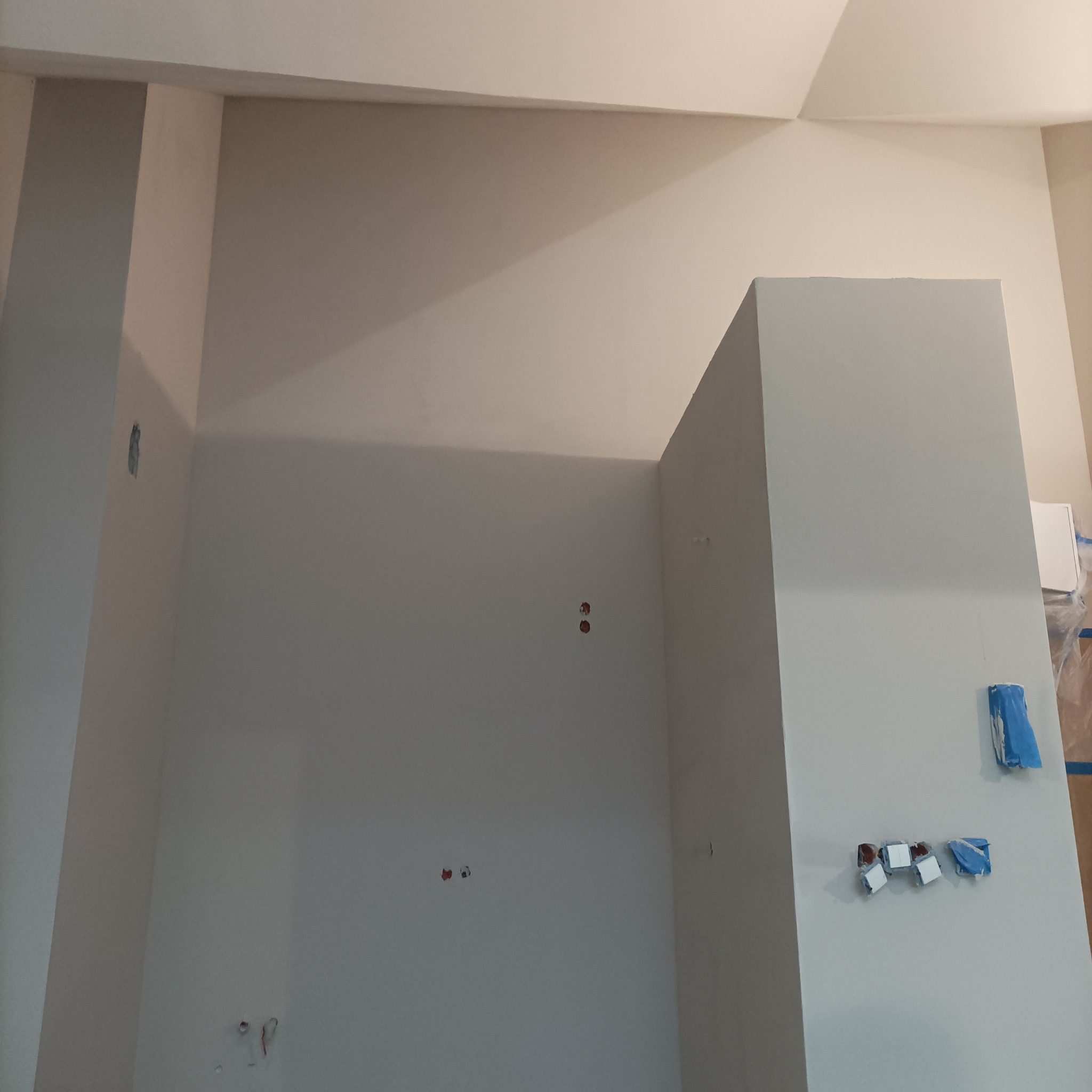 Świeżo pomalowane, jasnoszare wnętrze z widocznymi puszkami elektrycznymi i zabezpieczeniami taśmą malarską. Widok na ścianę i sufit pod kątem.