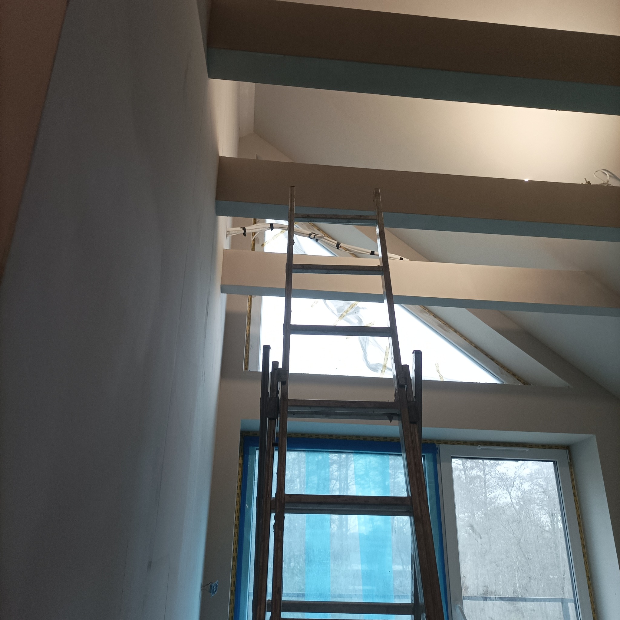Skośne okno dachowe zabezpieczone folią, widoczne zza drewnianej drabiny. Ściana w trakcie prac wykończeniowych, widoczne przewody elektryczne pod sufitem.