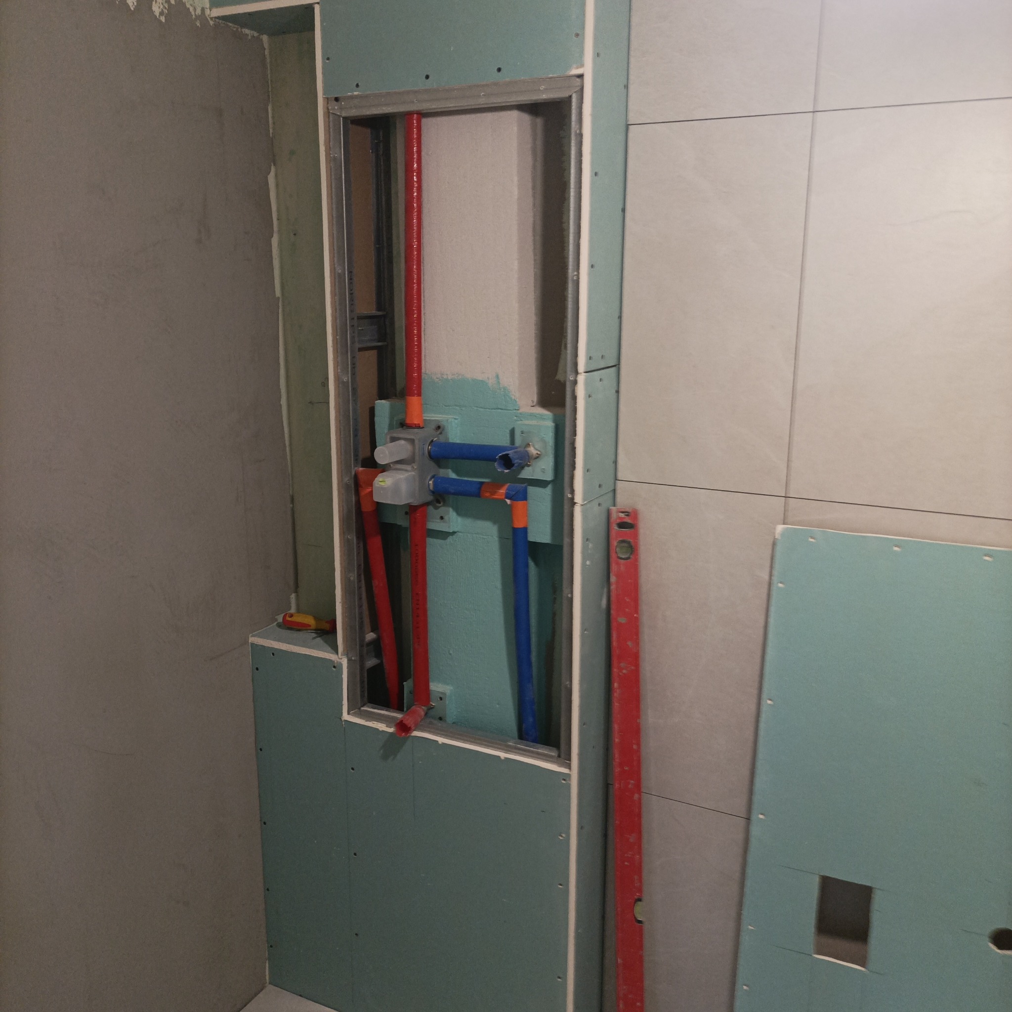 Narożnik łazienki w trakcie prac: widoczne płyty karton-gips, instalacja wodna z czerwonymi i niebieskimi rurami, poziomica. Etap przygotowania do wykończenia.