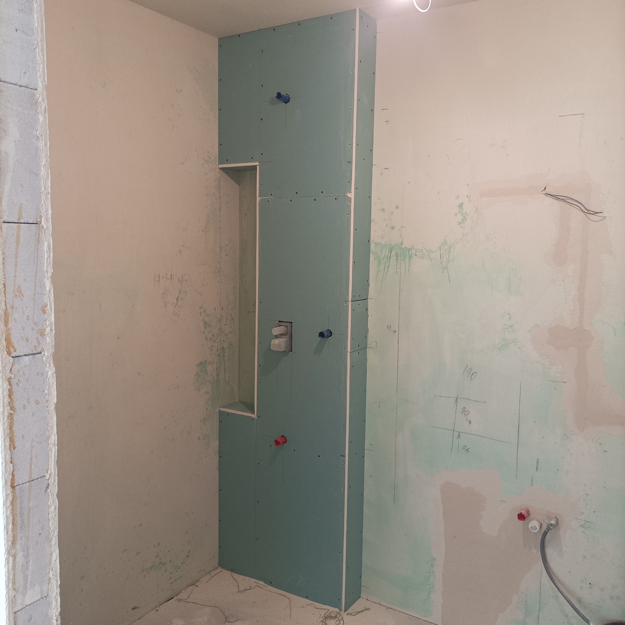 Narożna zabudowa z płyty karton-gips w surowej łazience, z widocznymi otworami na instalacje wodne i wnęką. Ściany z widocznymi śladami prac remontowych.