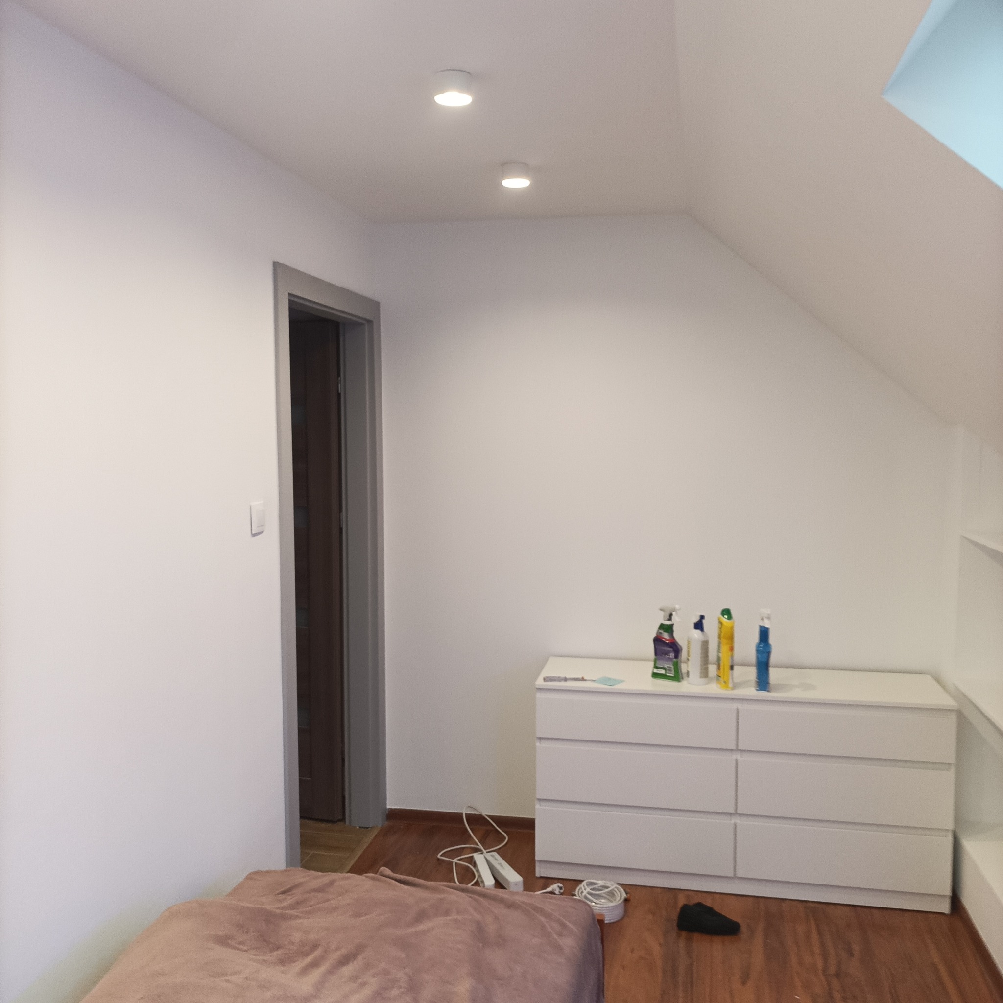 Pokój na poddaszu po remoncie: białe ściany, sufit ze światłami punktowymi, szare obramowanie drzwi, biała komoda z detergentami, drewniana podłoga i fragment łóżka.