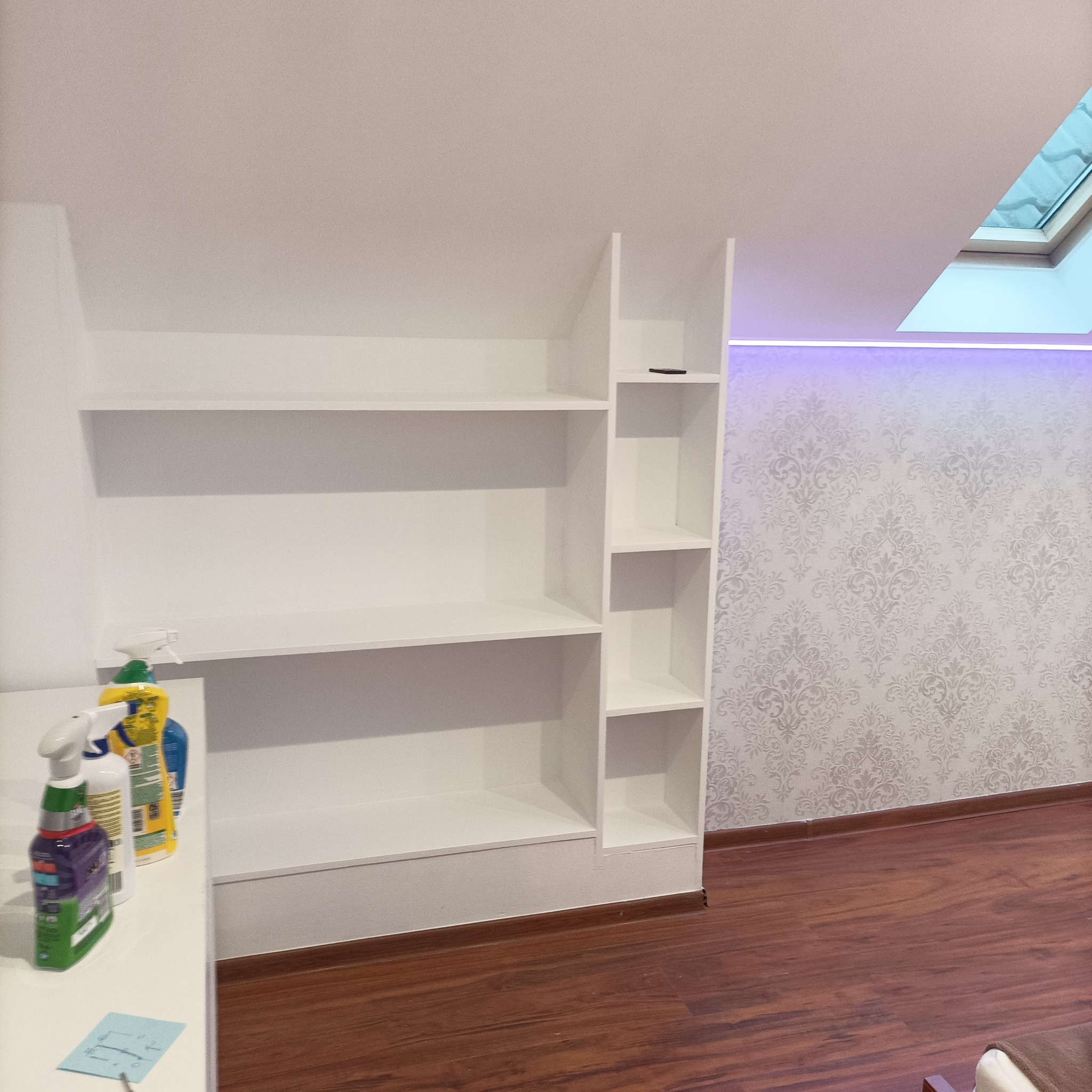 Biały regał na wymiar w pokoju na poddaszu, z widocznym oknem dachowym i dekoracyjną tapetą w tle. Na blacie obok regału stoją środki czystości.