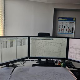 AW-instal Pracownia Projektowa Karolina Kruczkowska-Wężyk - Biuro projektowe: trzy monitory z planami instalacji, kalkulator, miarka, dokumenty. Widok zza biurka, światło dzienne z okna z żaluzjami.
