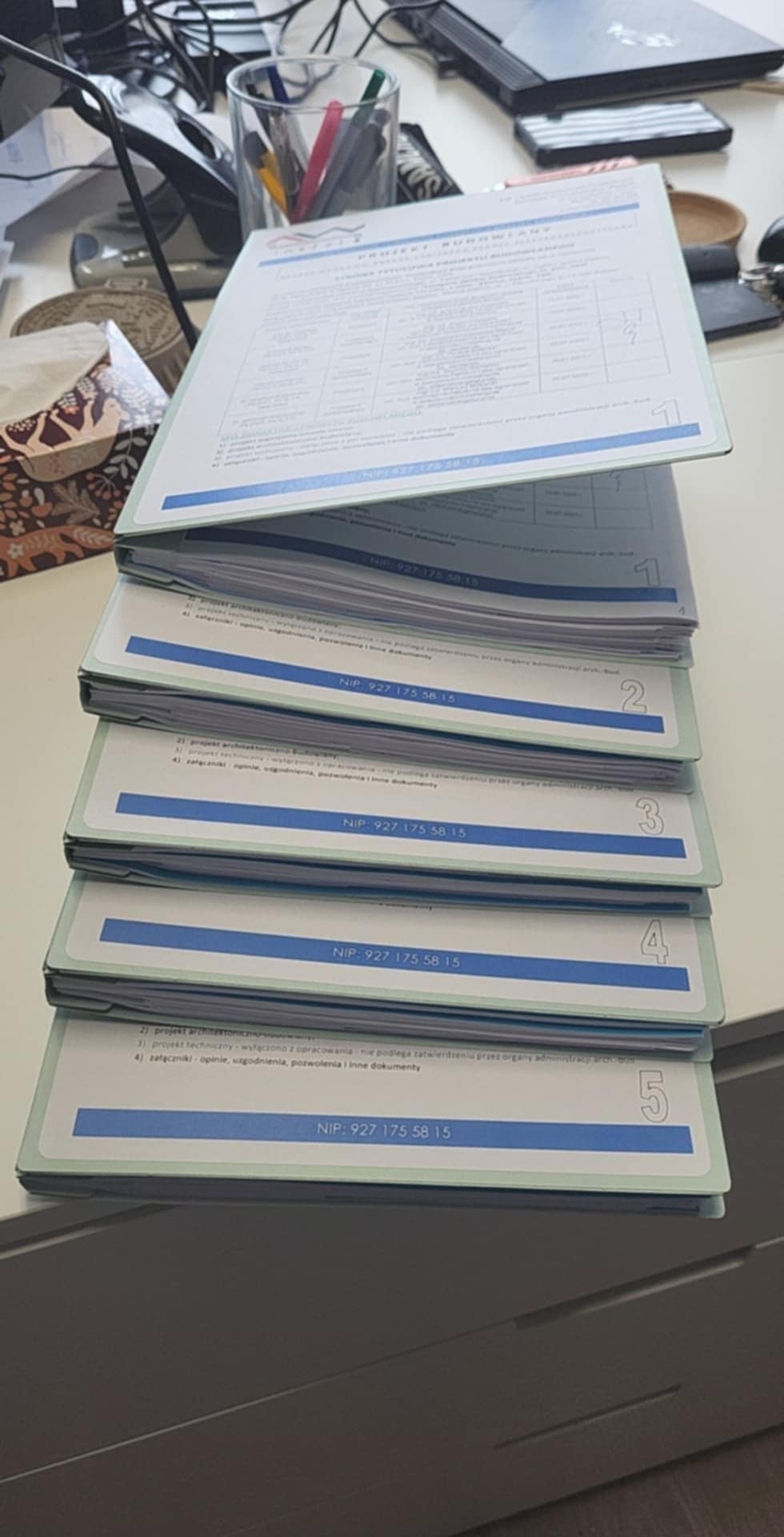 Stos dokumentów projektowych z numeracją 1-5, zawierających dane firmy (NIP). Dokumenty leżą na biurku, widoczne akcesoria biurowe i laptop w tle.