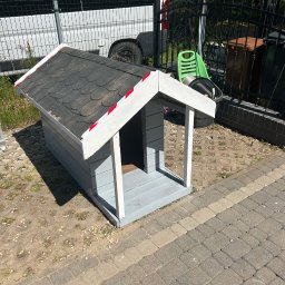 Bartosz Bojanowski - Szara buda dla psa z czarnym dachem, obok leży szczotka. W tle widoczny biały bus i ogrodzenie. Buda stoi na kostce brukowej i ziemi.