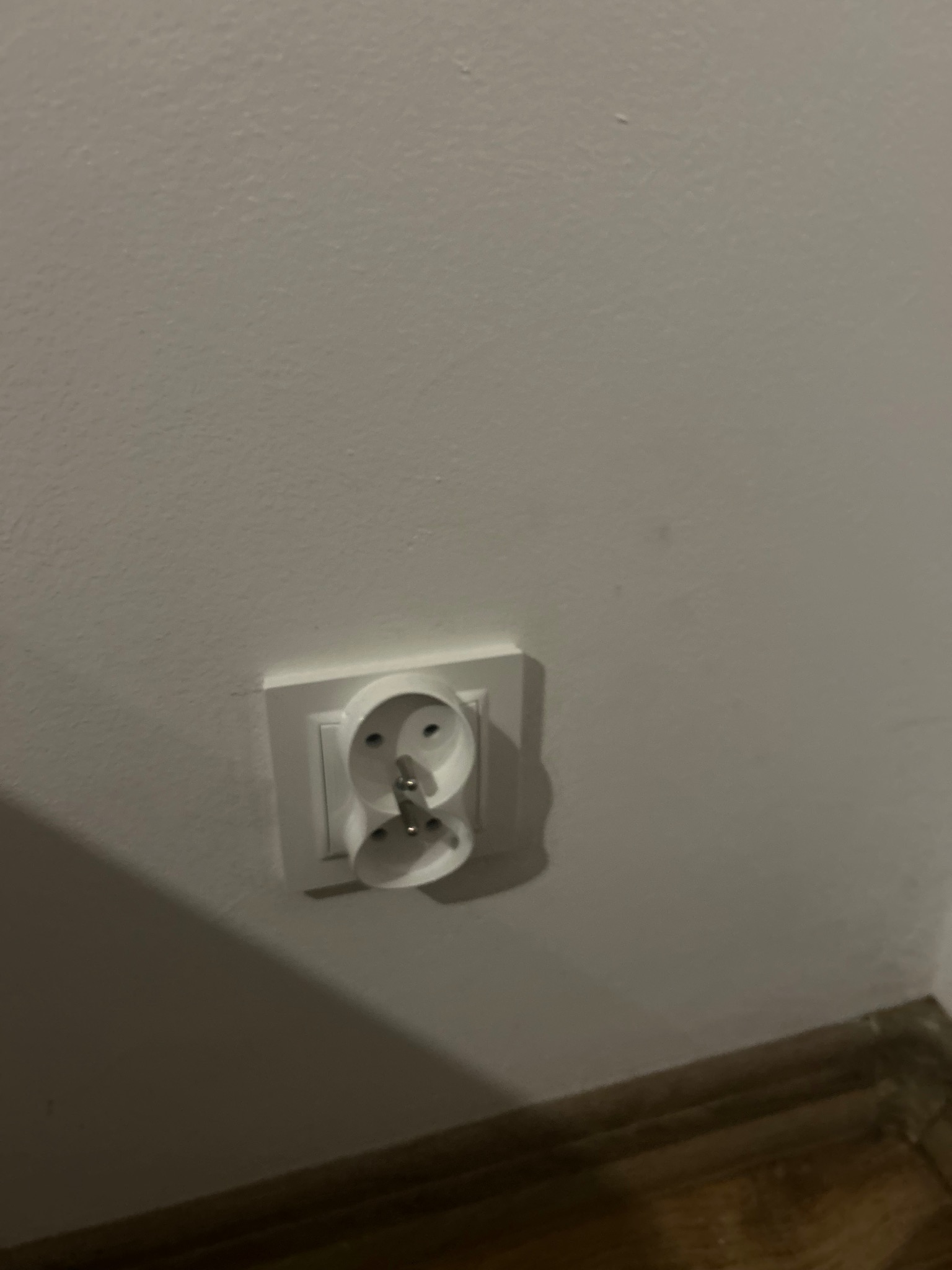 Białe gniazdko elektryczne z wtyczką na białej ścianie, widoczne drewniane wykończenie podłogi. Ujęcie z bliska, skupione na detalu instalacji elektrycznej.