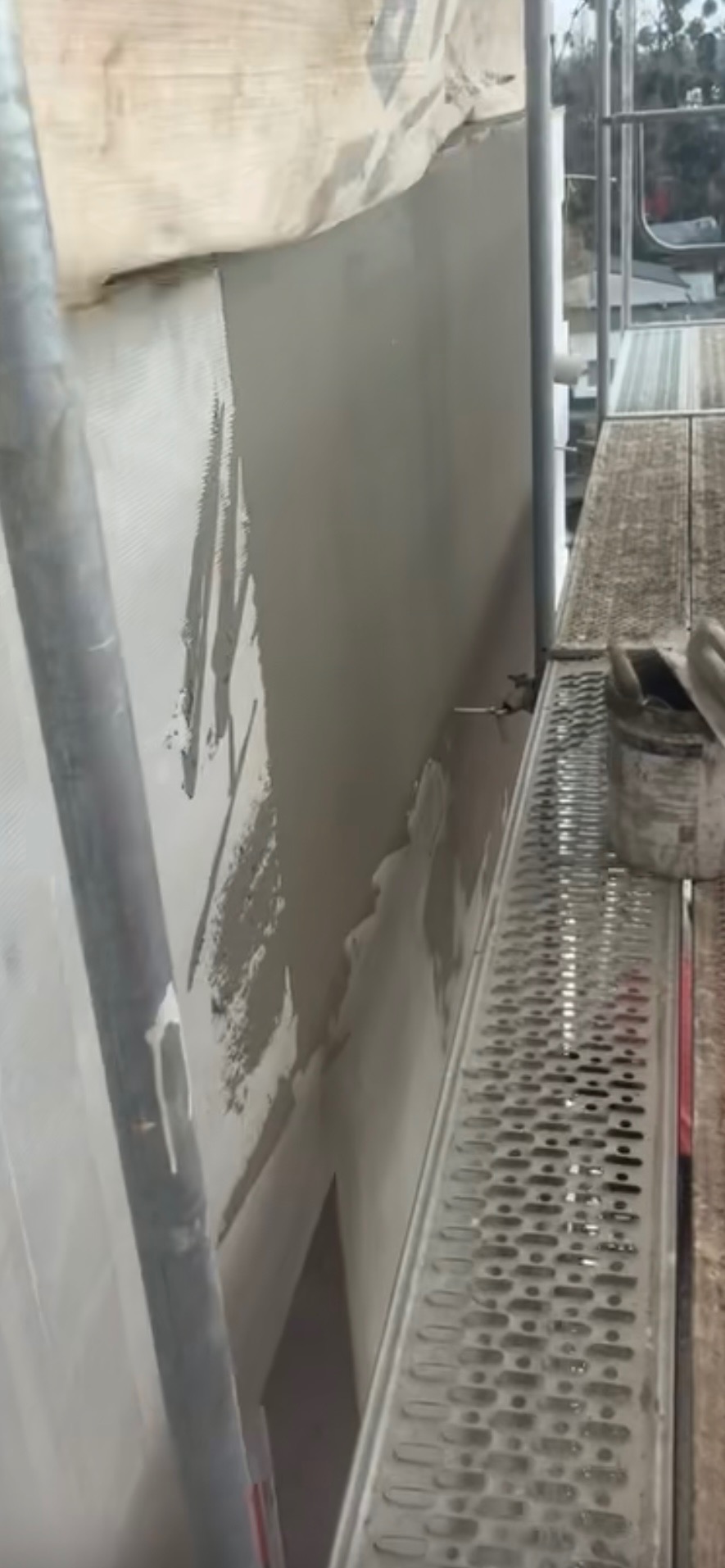 Ściana z nałożonym tynkiem, widoczna z rusztowania. Częściowo zakryta folią ochronną. Obok puszka z zaprawą. Ujęcie z dołu, podkreślające etap prac wykończeniowych.