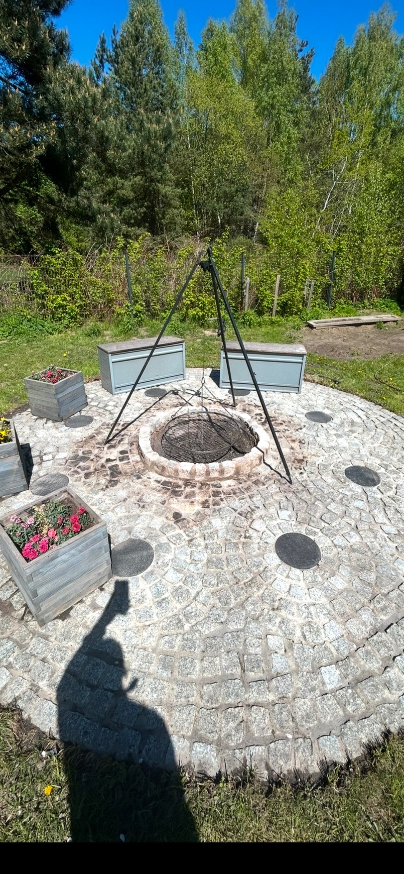Ognisko otoczone kamienną nawierzchnią z drewnianymi donicami i siedziskami, w tle zieleń drzew. Cień osoby robiącej zdjęcie w lewym dolnym rogu.