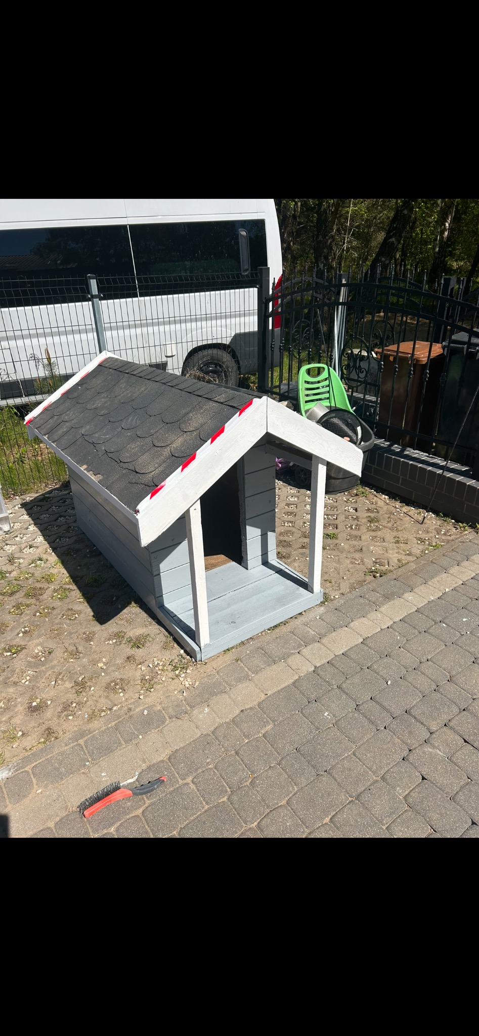 Szara buda dla psa z czarnym dachem, obok leży szczotka. W tle widoczny biały bus i ogrodzenie. Buda stoi na kostce brukowej i ziemi.