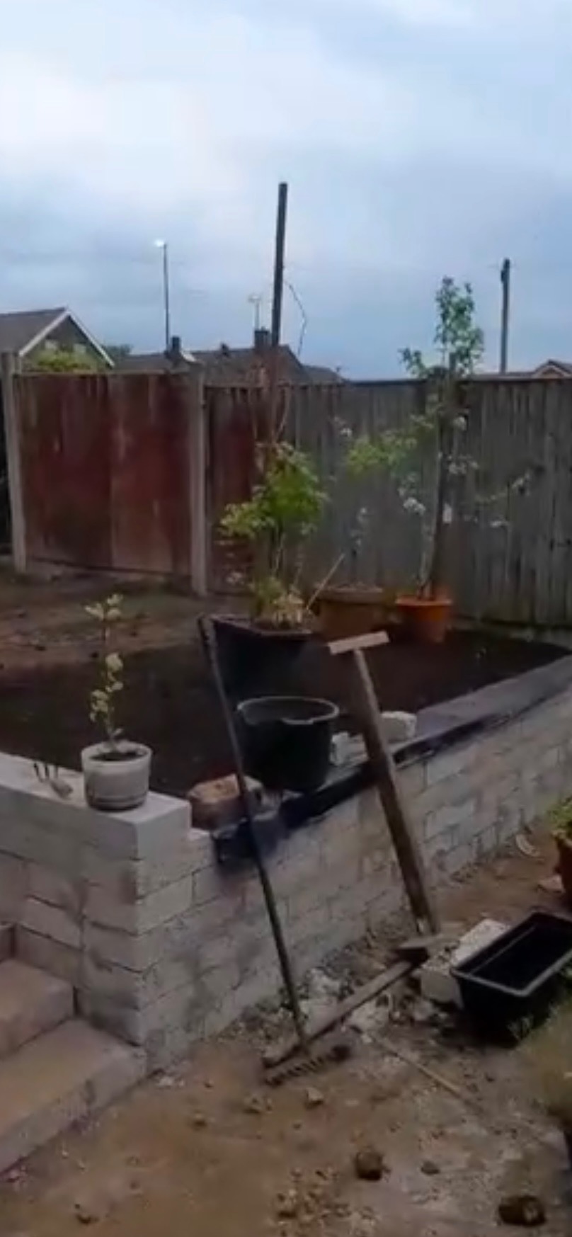 Budowa podwyższonej rabaty kwiatowej z cegły w ogrodzie w Gdańsku. Widoczne narzędzia ogrodnicze i donice z roślinami. Etap prac w toku, świeżo nasypana ziemia.