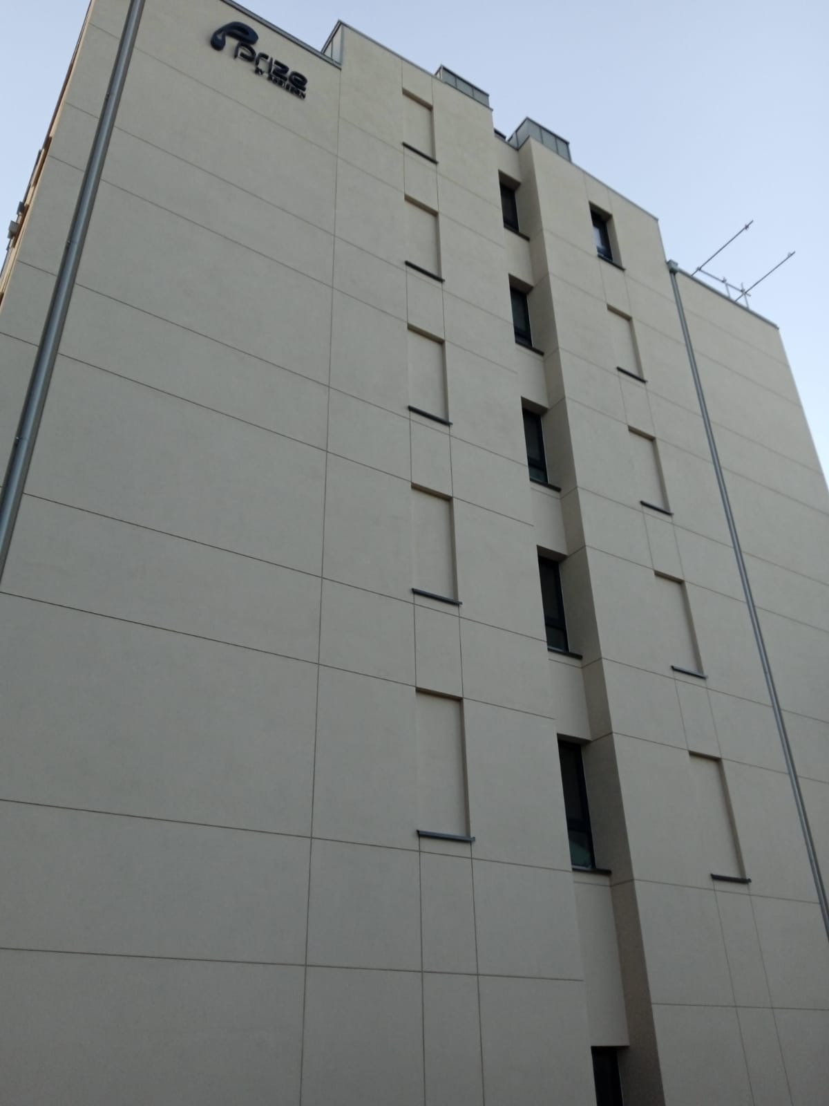 Wysoki budynek z beżową elewacją, widoczny z dołu pod kątem. Na szczycie logo firmy. Okna w pionowych rzędach. Minimalistyczny design.