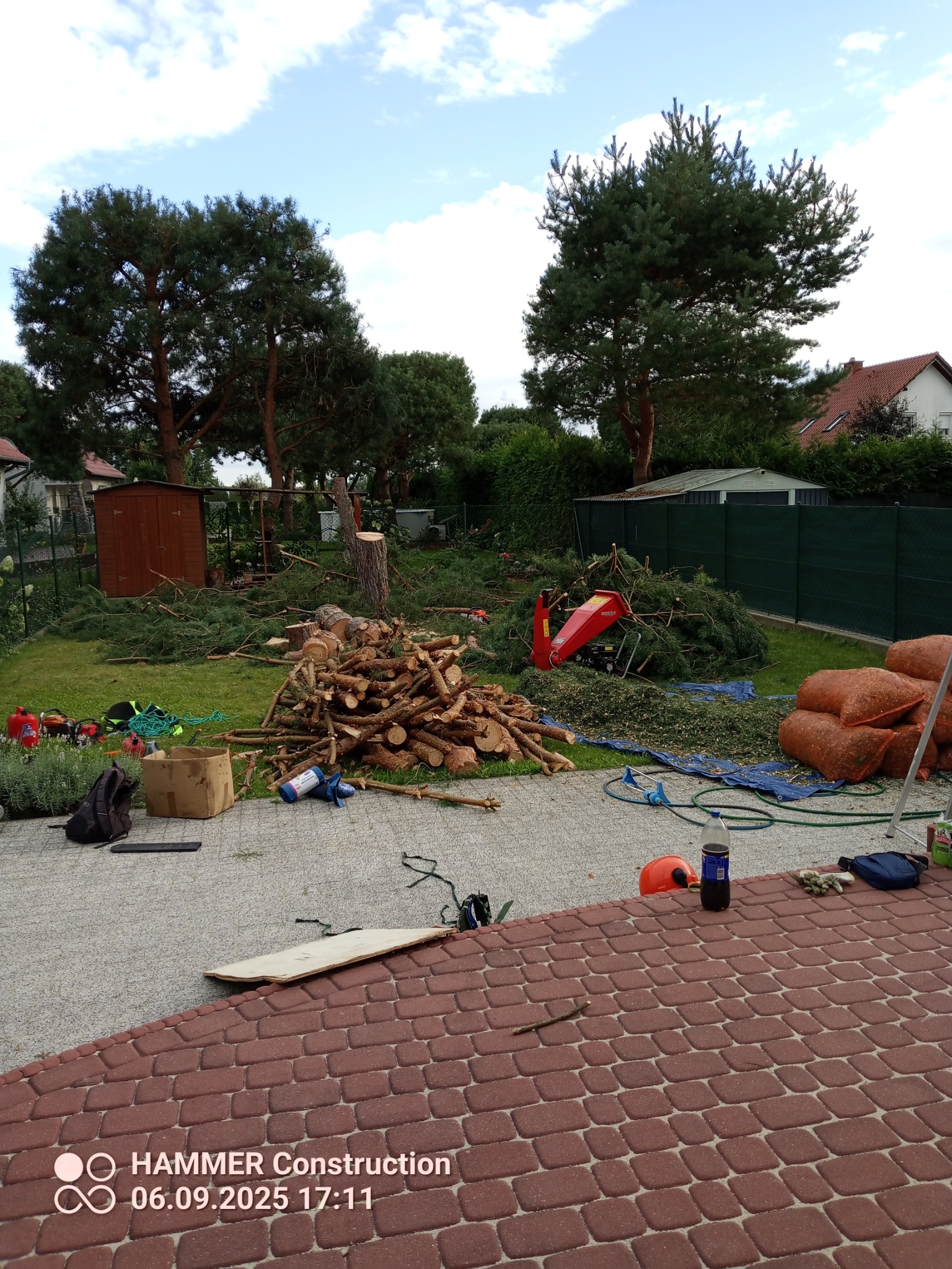 Teren po wycince drzew: pocięte pnie, gałęzie, rozdrabniarka, worki z odpadami. Widoczne narzędzia i akcesoria. Częściowo widoczny dach budynku w tle.