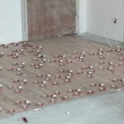 Dar-bud usługi ogólno budowlane - Układanie płytek imitujących drewno z użyciem klipsów poziomujących. Widoczny fragment ściany i drzwi. Na pierwszym planie wiadro z zaprawą.