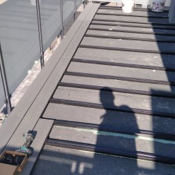 Dar-bud usługi ogólno budowlane - Montaż deski kompozytowej na balkonie z szarą balustradą. Widoczne narzędzia i cień osoby wykonującej prace. Etap budowy tarasu na dachu budynku.