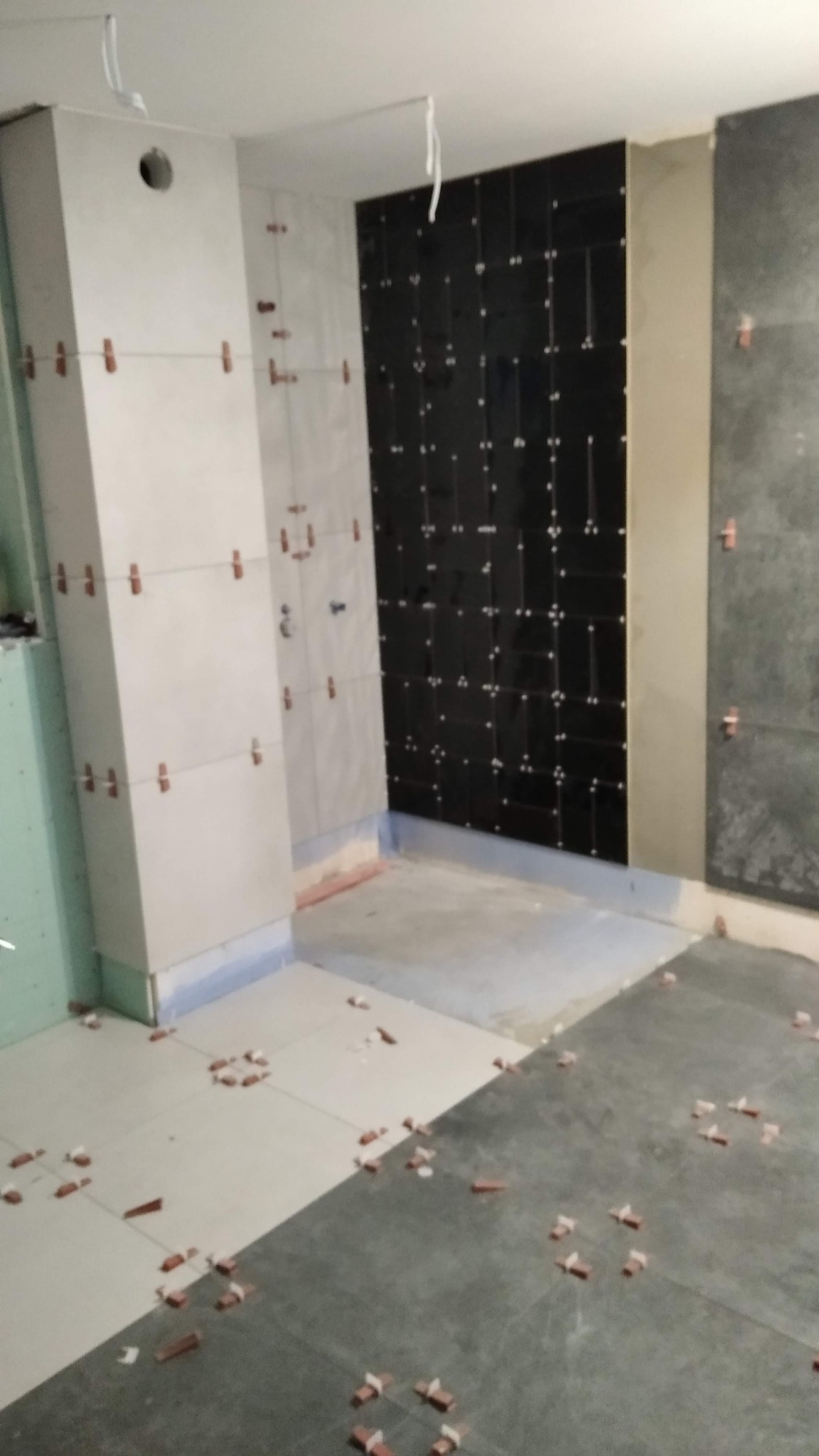 Wnętrze łazienki w trakcie remontu: częściowo ułożone płytki na ścianach i podłodze, widoczne krzyżyki dystansowe. Prace wykończeniowe w toku.