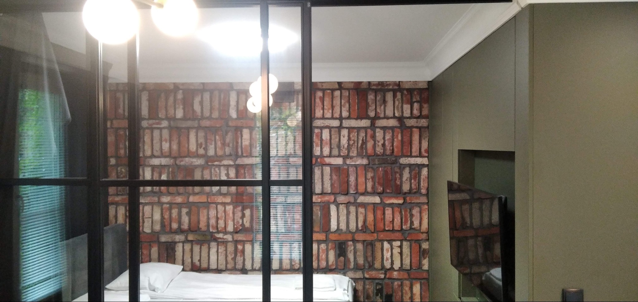 Loftowa sypialnia za szklaną ścianą z czarnymi szprosami. Ceglana tapeta na ścianie, nowoczesne oświetlenie i minimalistyczny wystrój.