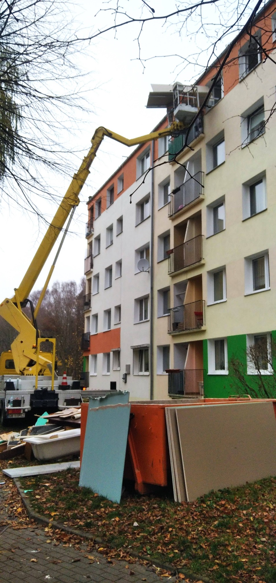 Żółty podnośnik koszowy przy bloku, płyty gipsowe i gruz na ziemi. Remont elewacji budynku wielorodzinnego w trakcie prac.