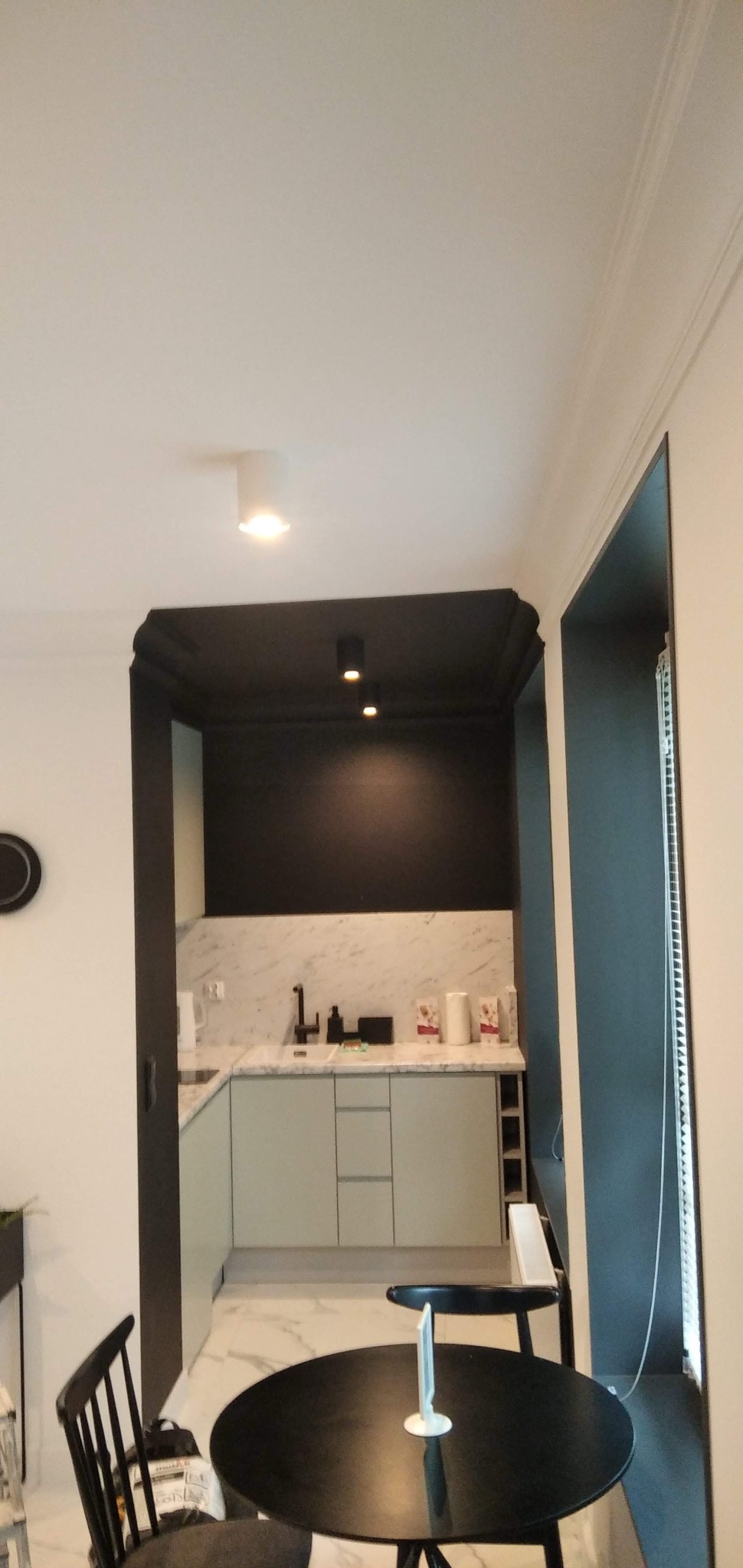 Nowoczesna kuchnia z czarnym sufitem i marmurowym blatem, oświetlona punktowymi lampami. Widoczny stół z krzesłami i fragment okna z żaluzjami.