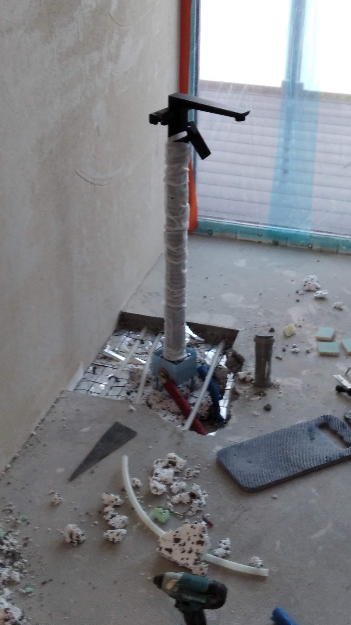 Czarna bateria umywalkowa na rurze w folii ochronnej, wystająca z podłogi w trakcie remontu łazienki. Widoczne rury, narzędzia i gruz.