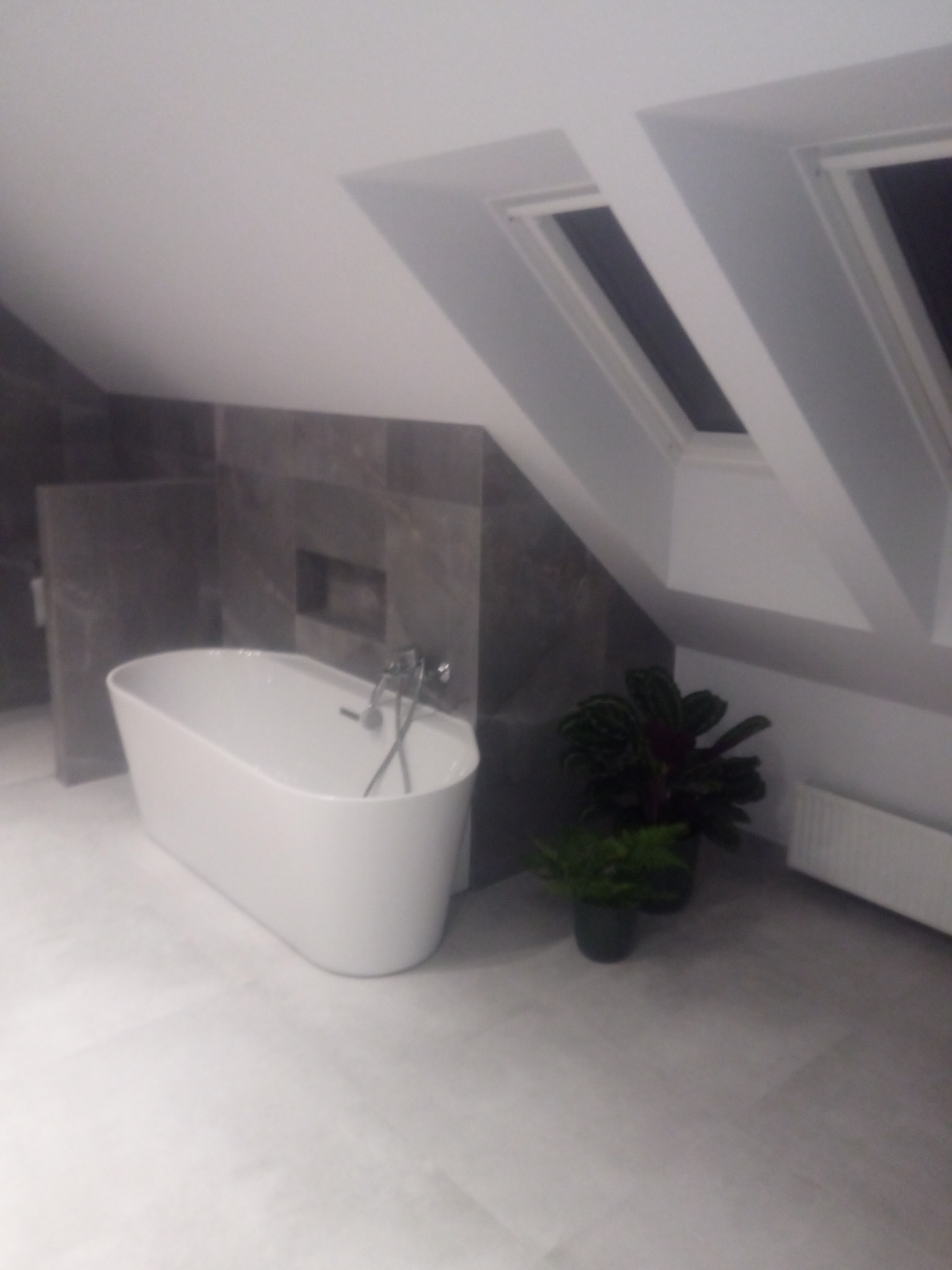 Nowoczesna łazienka na poddaszu z wolnostojącą wanną, szarą okładziną ścienną i oknami dachowymi. Uzupełniona o rośliny doniczkowe.