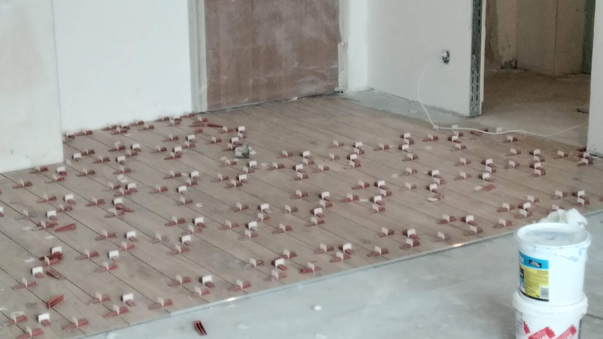 Układanie płytek imitujących drewno z użyciem klipsów poziomujących. Widoczny fragment ściany i drzwi. Na pierwszym planie wiadro z zaprawą.