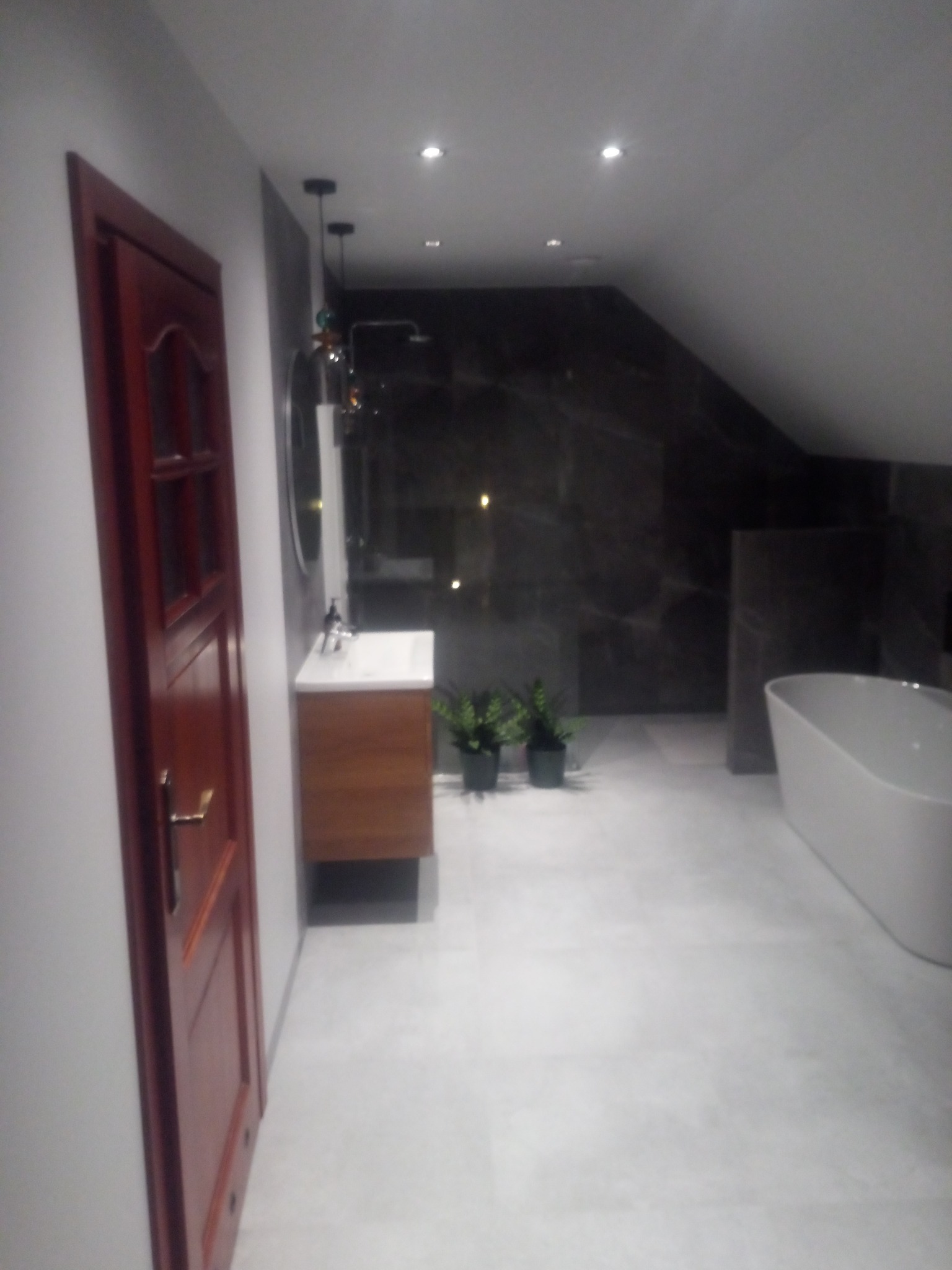 Nowoczesna łazienka na poddaszu z drewnianą szafką pod umywalką, marmurowymi ścianami i wolnostojącą wanną. Ciemne wykończenie i punktowe oświetlenie.