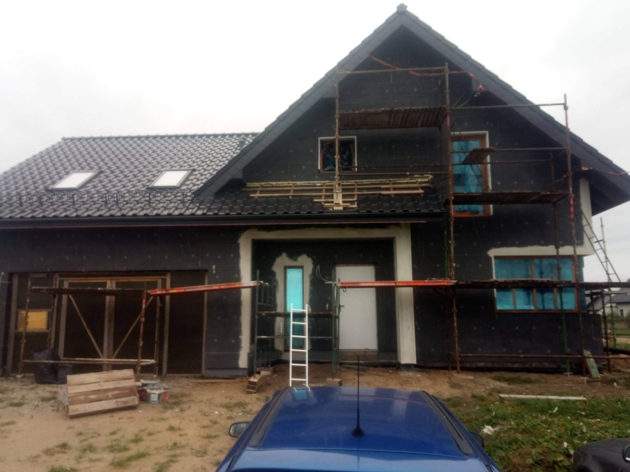 Dom jednorodzinny w trakcie ocieplania, widoczne rusztowania i materiały izolacyjne. Okna zabezpieczone folią, przygotowania do elewacji.