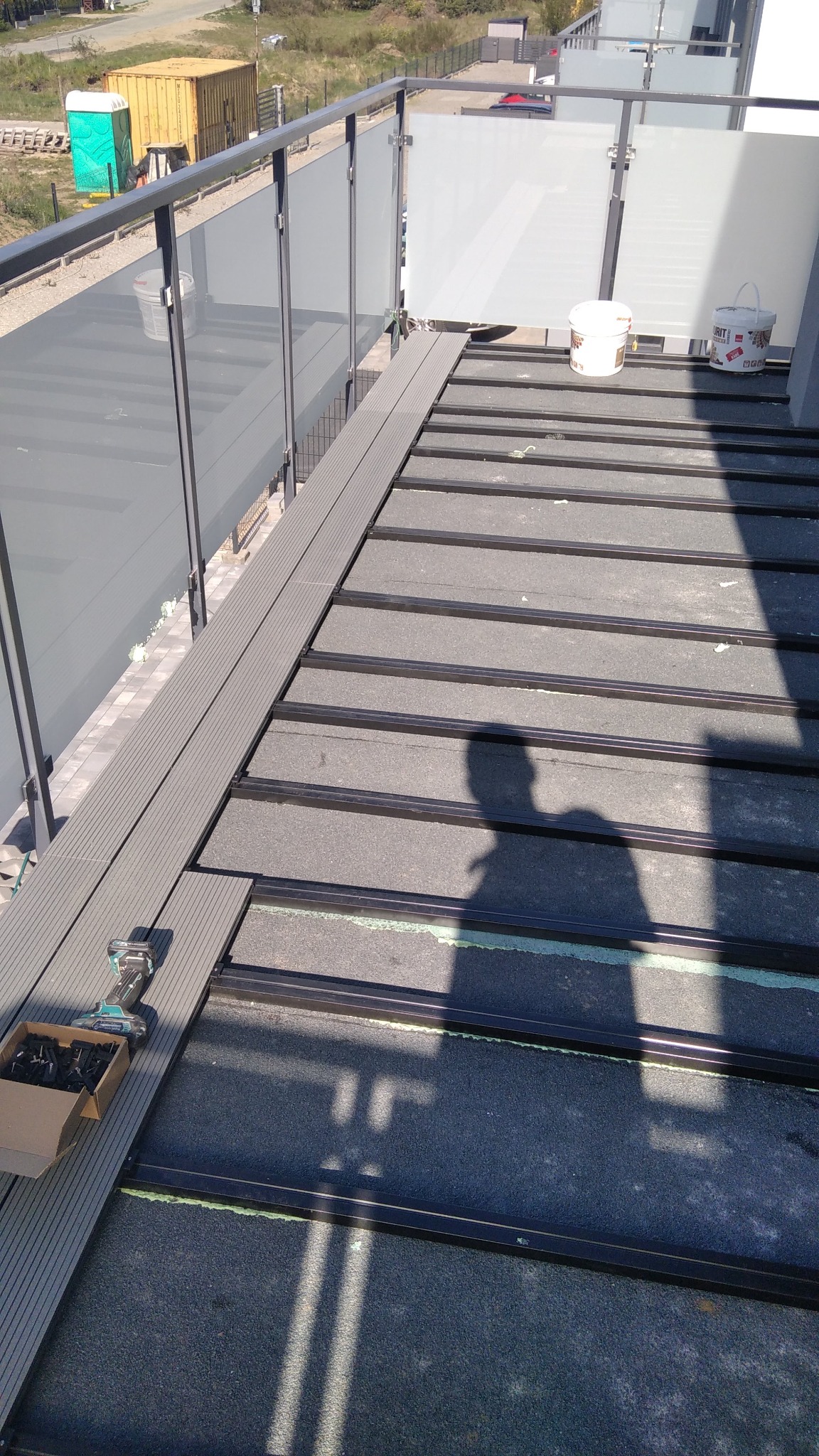 Montaż deski kompozytowej na balkonie z szarą balustradą. Widoczne narzędzia i cień osoby wykonującej prace. Etap budowy tarasu na dachu budynku.