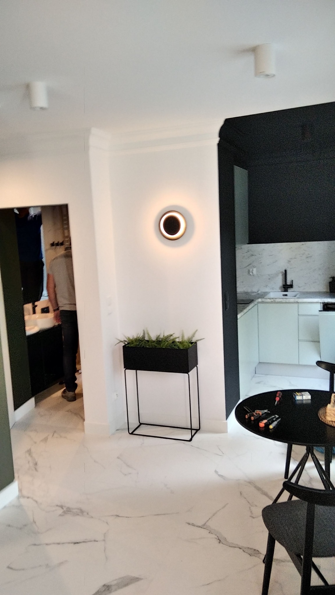 Nowoczesne wnętrze po remoncie: białe ściany, marmurowe płytki podłogowe, czarne akcenty, oświetlenie punktowe i dekoracje roślinne. Widok na odnowioną kuchnię.