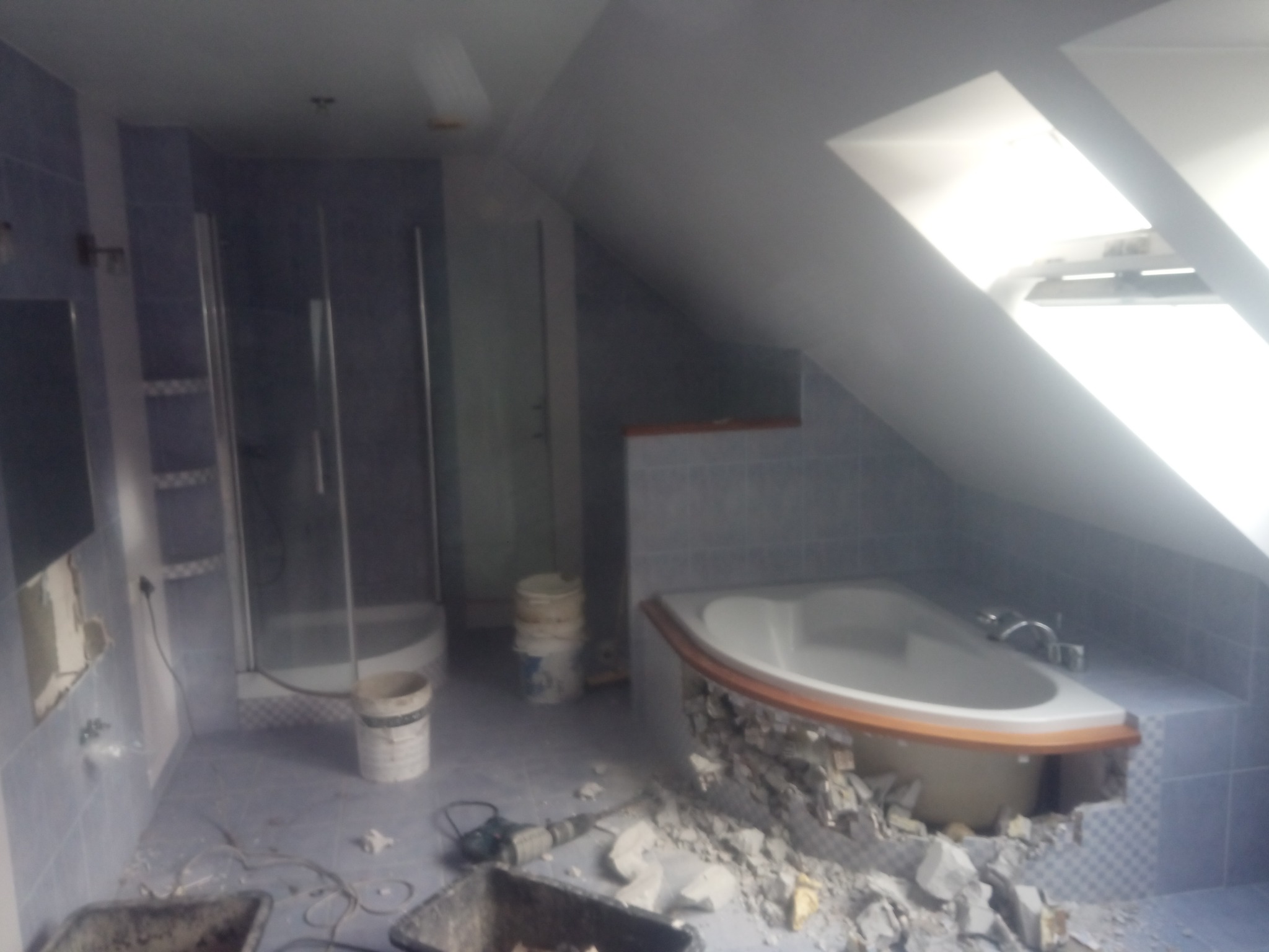 Demontaż obudowy wanny w łazience na poddaszu. Widoczne gruz, narzędzia i fragment kabiny prysznicowej. Etap prac remontowych w pomieszczeniu z oknem dachowym.