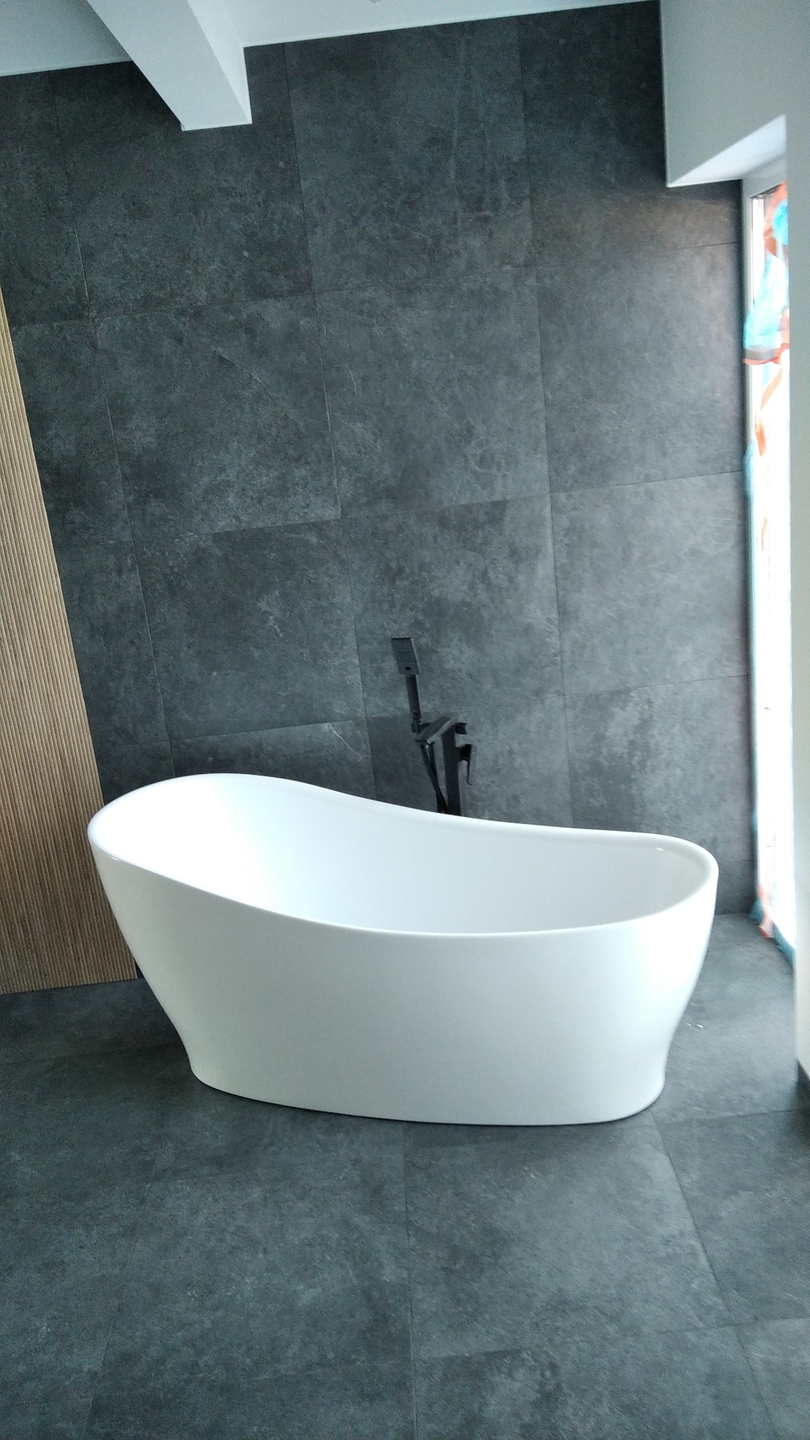 Nowoczesna, wolnostojąca wanna w łazience z szarymi płytkami i czarną armaturą. Minimalistyczny design, jasne wnętrze z akcentem drewnianej ściany.