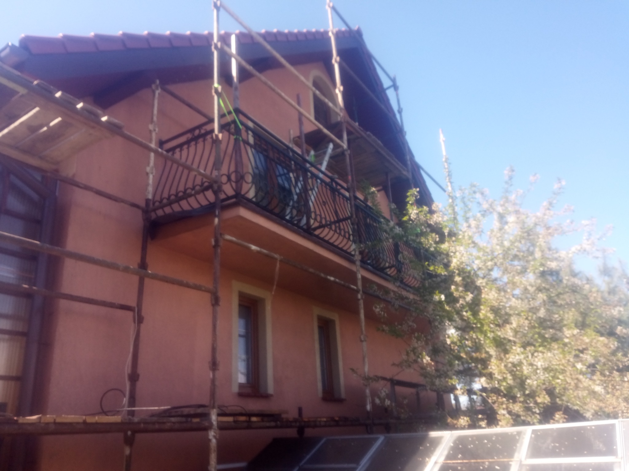 Dom jednorodzinny w trakcie malowania elewacji, otoczony rusztowaniami. Widoczny balkon z ozdobną balustradą i kwitnące drzewo w tle. Ujęcie z dołu.