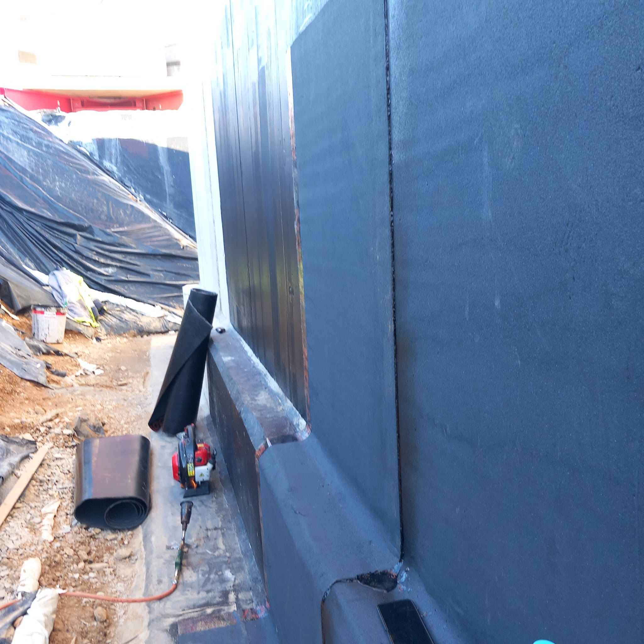 Hydroizolacja pionowa fundamentów: czarna powłoka ochronna na ścianie, obok rozłożona papa bitumiczna i narzędzia. Widoczne zabezpieczenia wykopu.