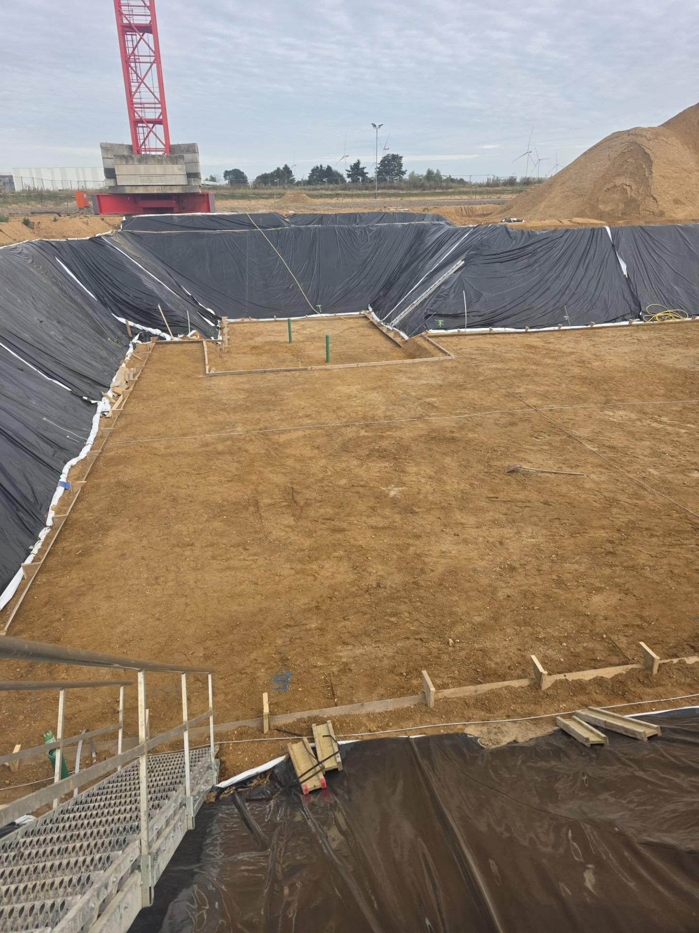 Przygotowanie terenu pod budowę: wykop wyłożony czarną folią, drewniane szalunki, widoczny żuraw budowlany i hałda piasku w tle. Prace ziemne w toku.