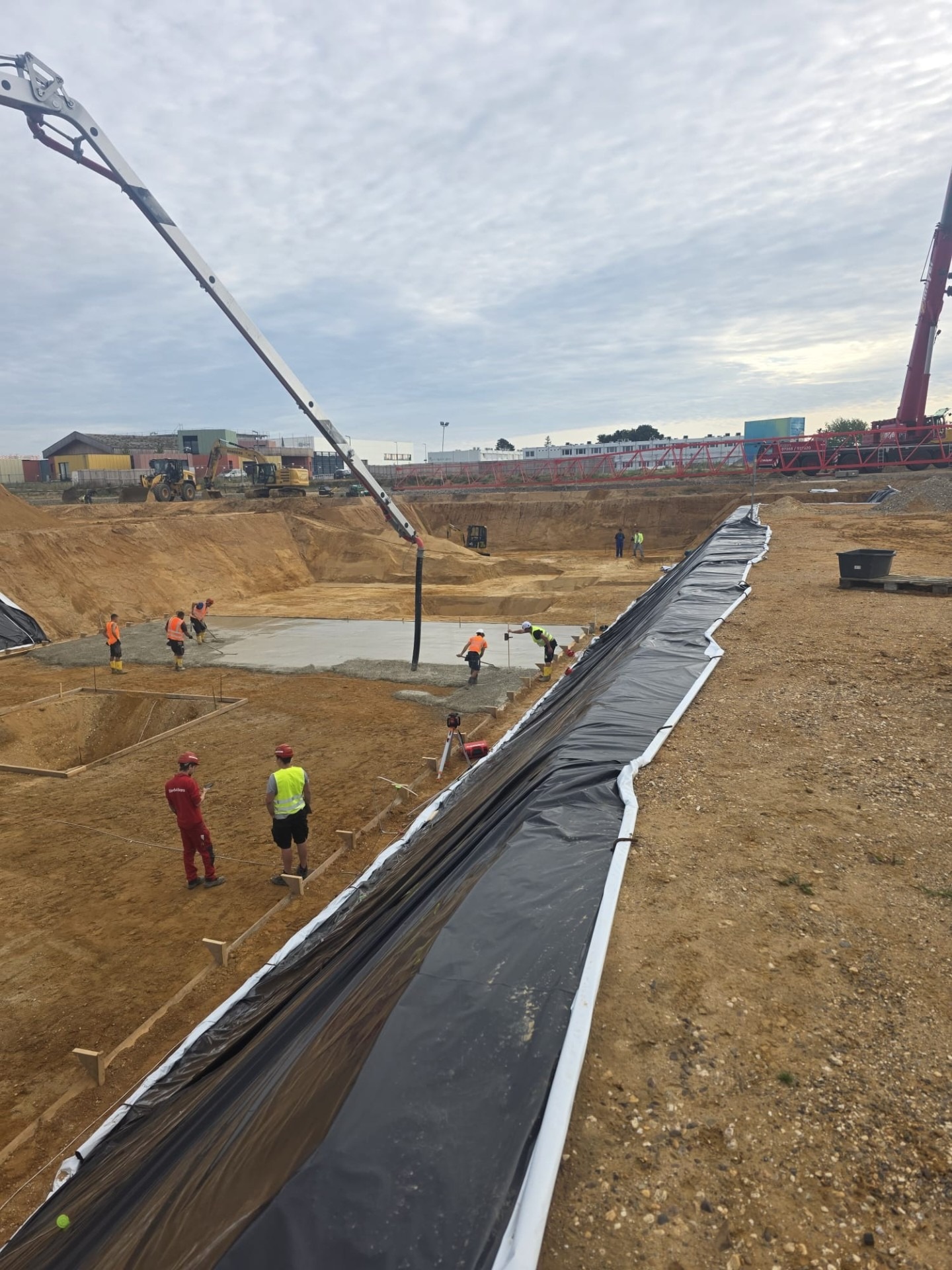 Wylewanie fundamentów na budowie z użyciem pompy do betonu. Widoczni pracownicy w odblaskowych kamizelkach i kaskach. W tle koparki i dźwig.
