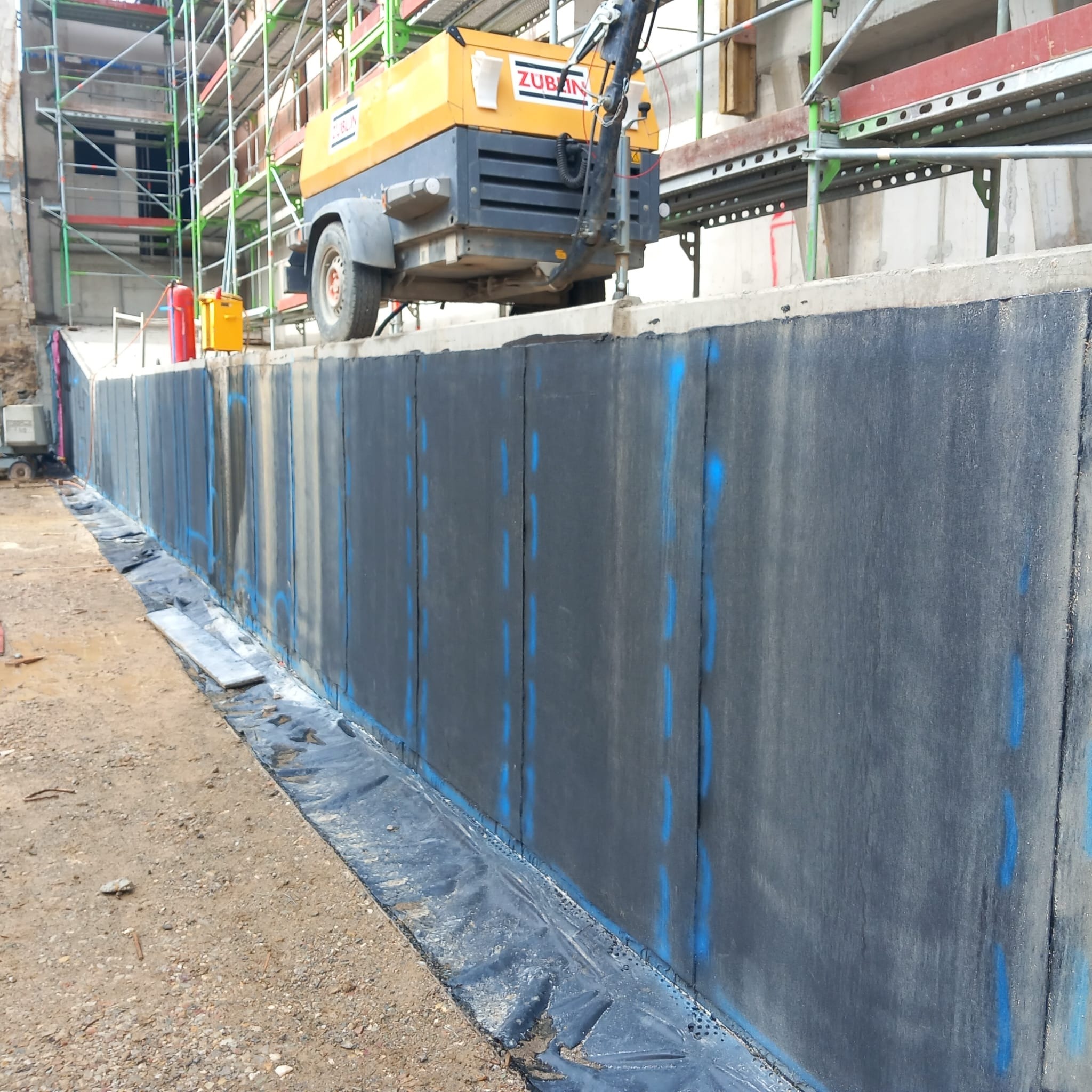 Hydroizolacja pionowa fundamentów z zaznaczonymi punktami mocowania, widoczna folia ochronna i rusztowania w tle. Prace budowlane w toku.