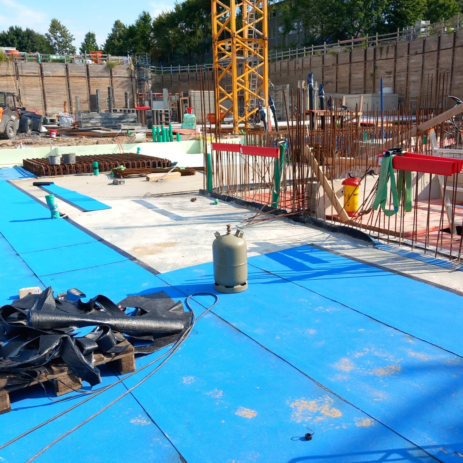 Niebieska izolacja na budowie z widoczną konstrukcją zbrojeniową, butlą gazową i żurawiem w tle. Elementy przygotowane do wylania posadzki.