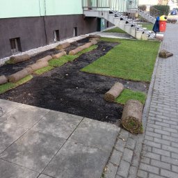 F.H.U Dębowa Kraina - Układanie trawy z rolki na przygotowanym podłożu, widoczne fragmenty ułożonej trawy i zwinięte rolki, tło stanowi budynek z zewnętrznymi schodami.