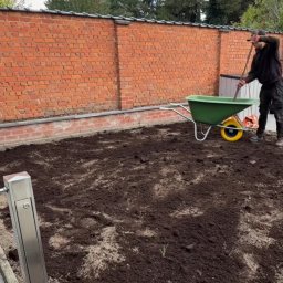 F.H.U Dębowa Kraina - Przygotowanie gruntu pod trawnik: mężczyzna z taczką i narzędziem wyrównuje ziemię na działce, widoczny ceglany mur i fragment instalacji nawadniającej.